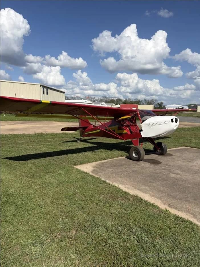 A red and yellow ultralight aircraft with a white nose cowl parked on grass next to a concrete pad under a blue sky with clouds.