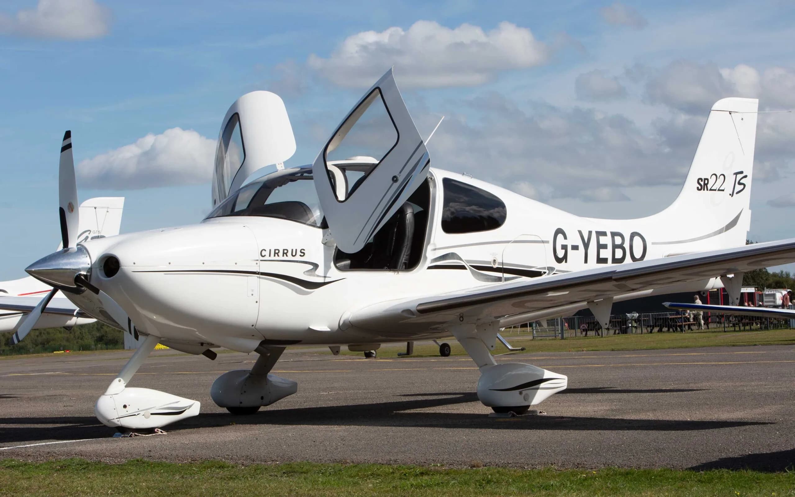 A white Cirrus SR22 TSi aircraft with black and grey stripes, registration G-YEBO, parked on an airfield with the right gull-wing door open.