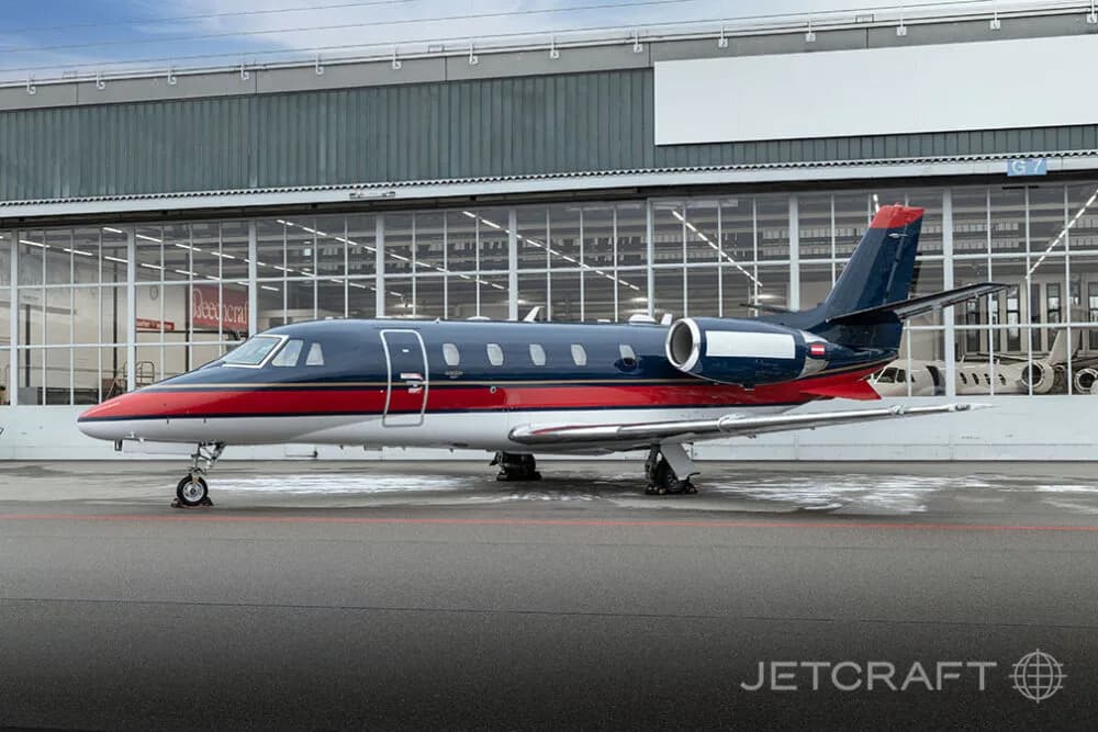 A dark blue, red, and white business jet parked on an asphalt apron with a hangar in the background.