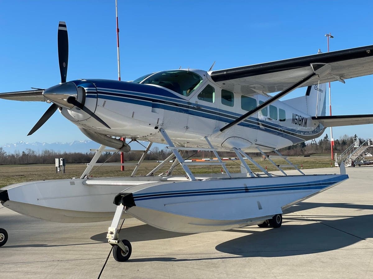A front-quarter view of a white and blue Cessna Caravan on floats, showing the propeller and front of the aircraft.