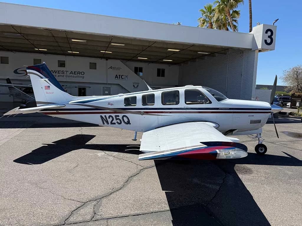 A white Beechcraft Bonanza A36, registration N25Q, with blue and red stripes, parked on a tarmac next to a hangar.