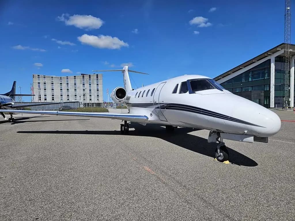 A white business jet with black stripes parked on an airfield with modern buildings in the background.