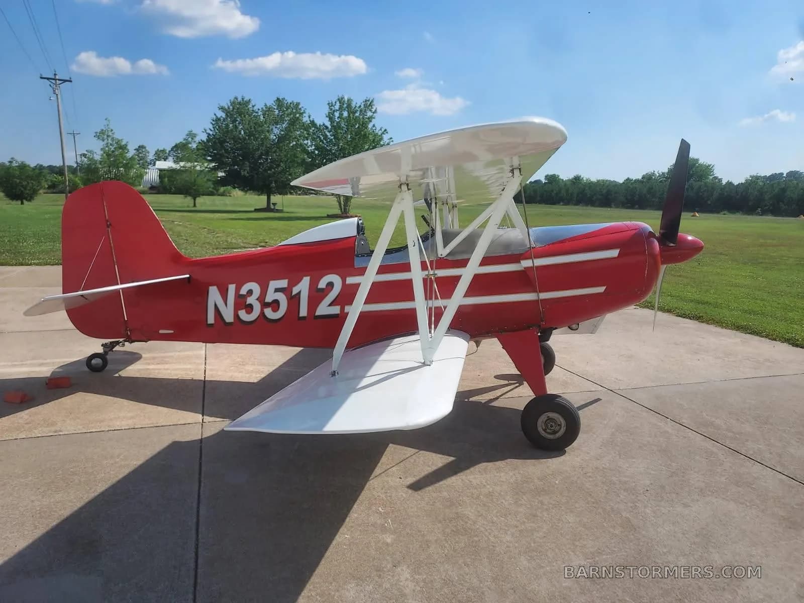 A red biplane with white wings and N3512 on the fuselage, viewed from the side on a concrete pad with a grassy field behind it.