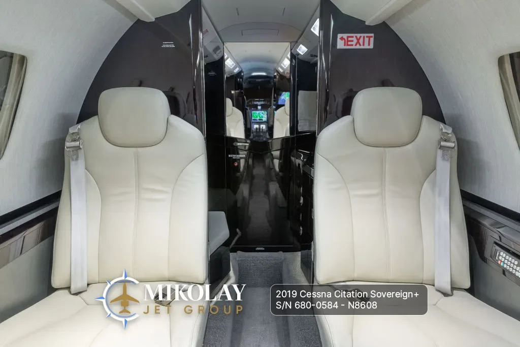 A view from the rear of the aircraft cabin, showing two light-colored leather seats facing forward and the galley area.