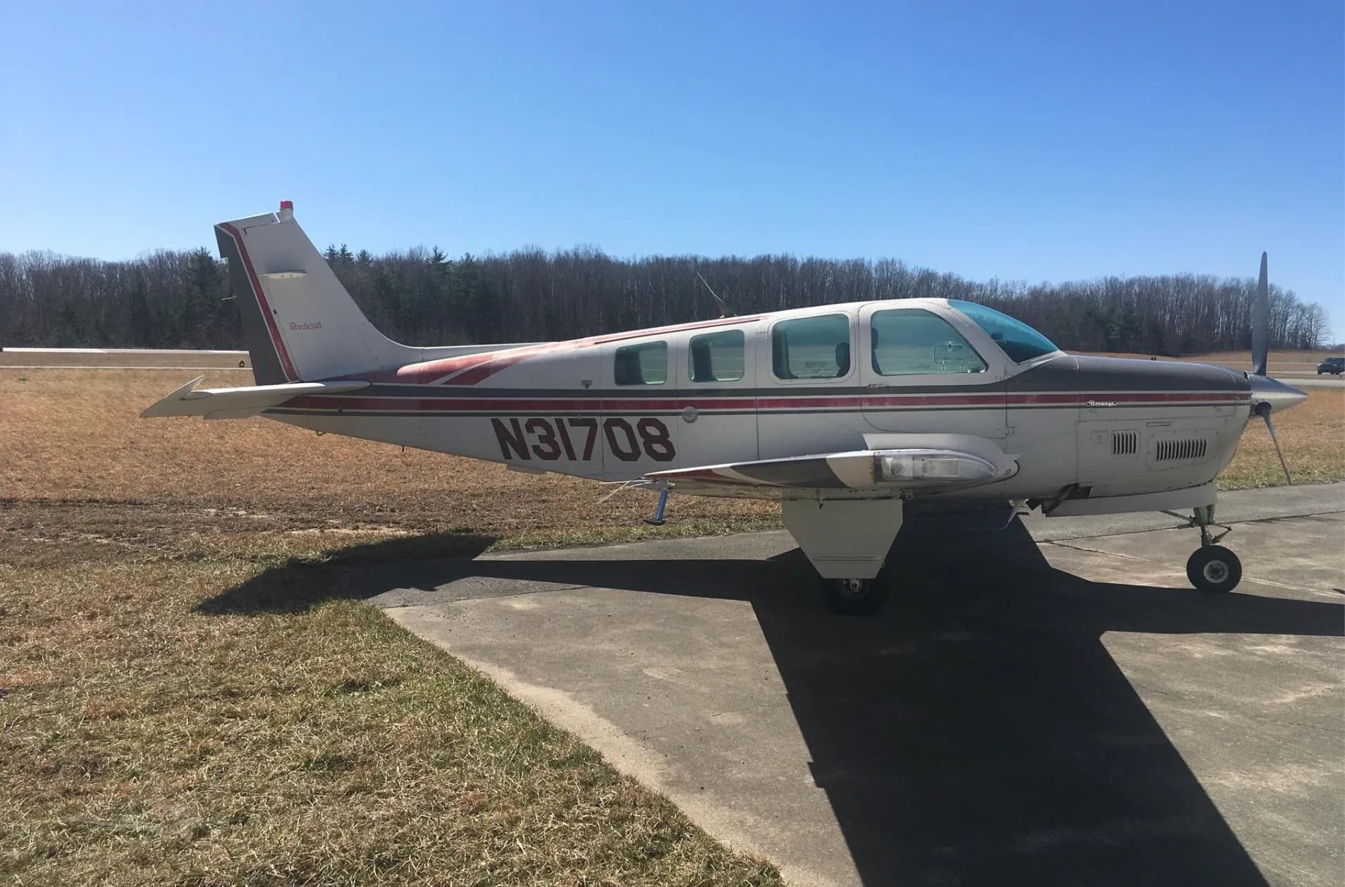 A side view of a white and red Beechcraft Bonanza N31708 parked on an asphalt ramp with a distant road and trees in the background.