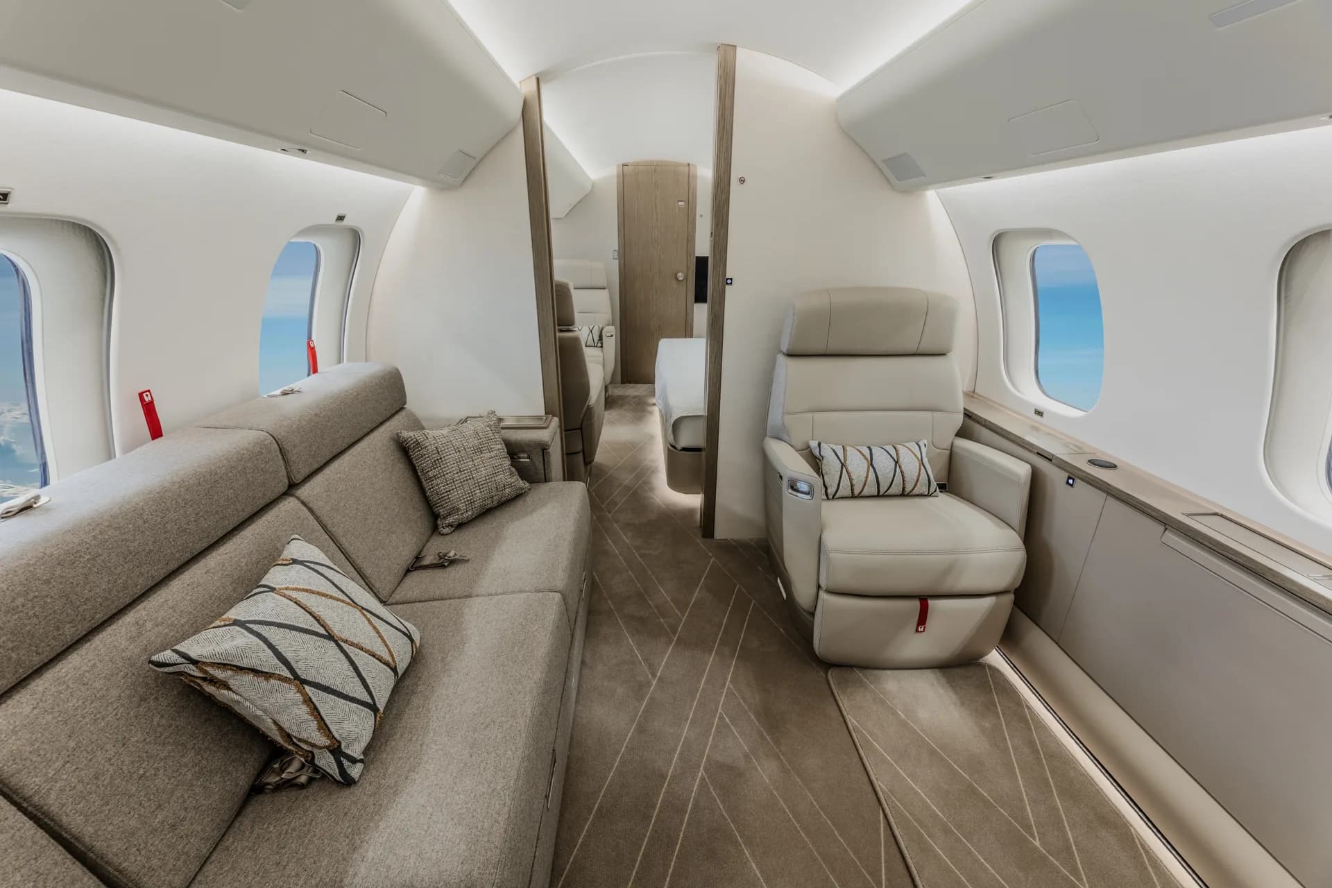A view inside a private jet cabin showing a long grey sofa with pillows and a single cream leather seat.
