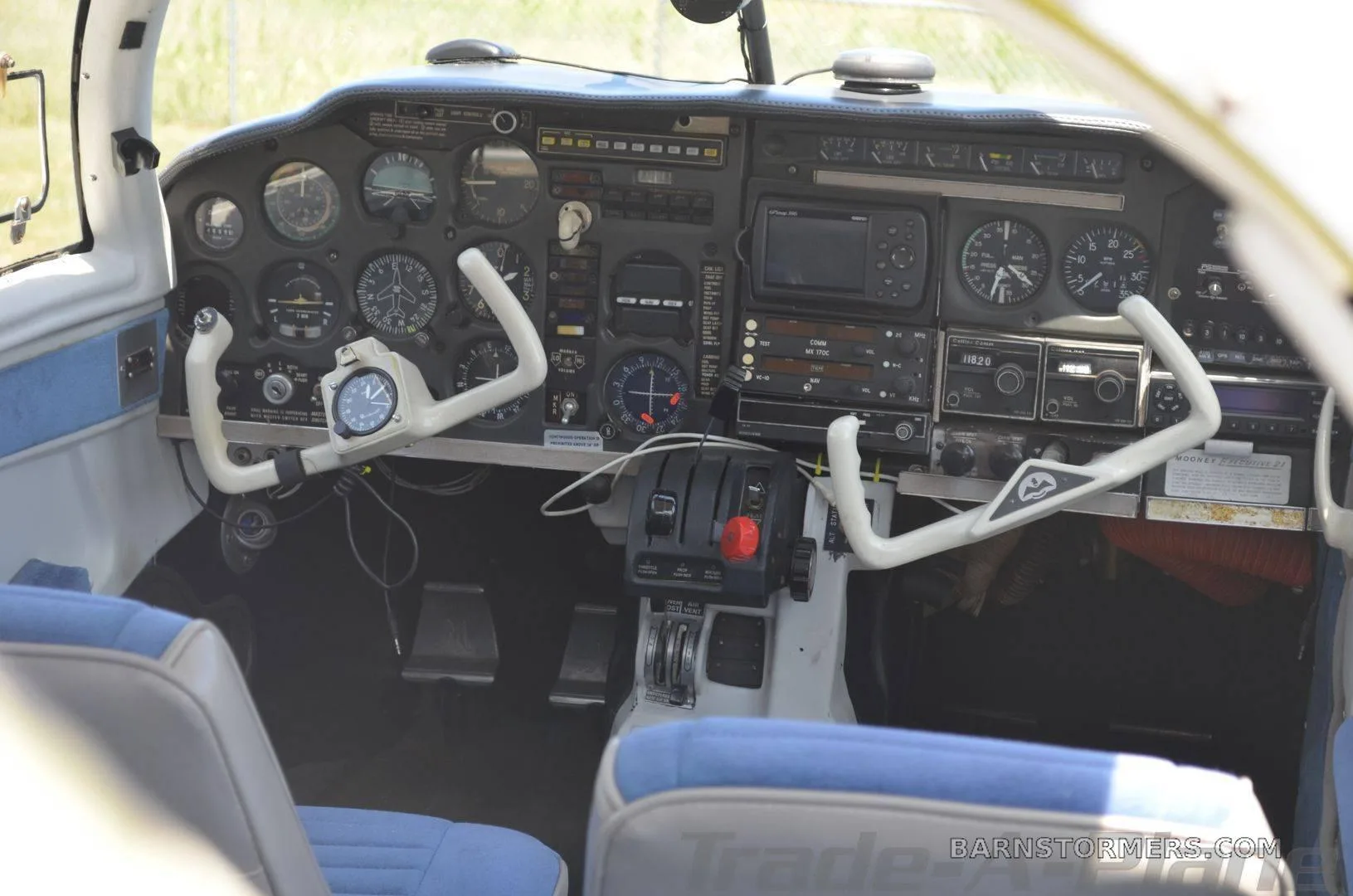 The cockpit of a small aircraft features a detailed instrument panel with dual control yokes and blue fabric seats.