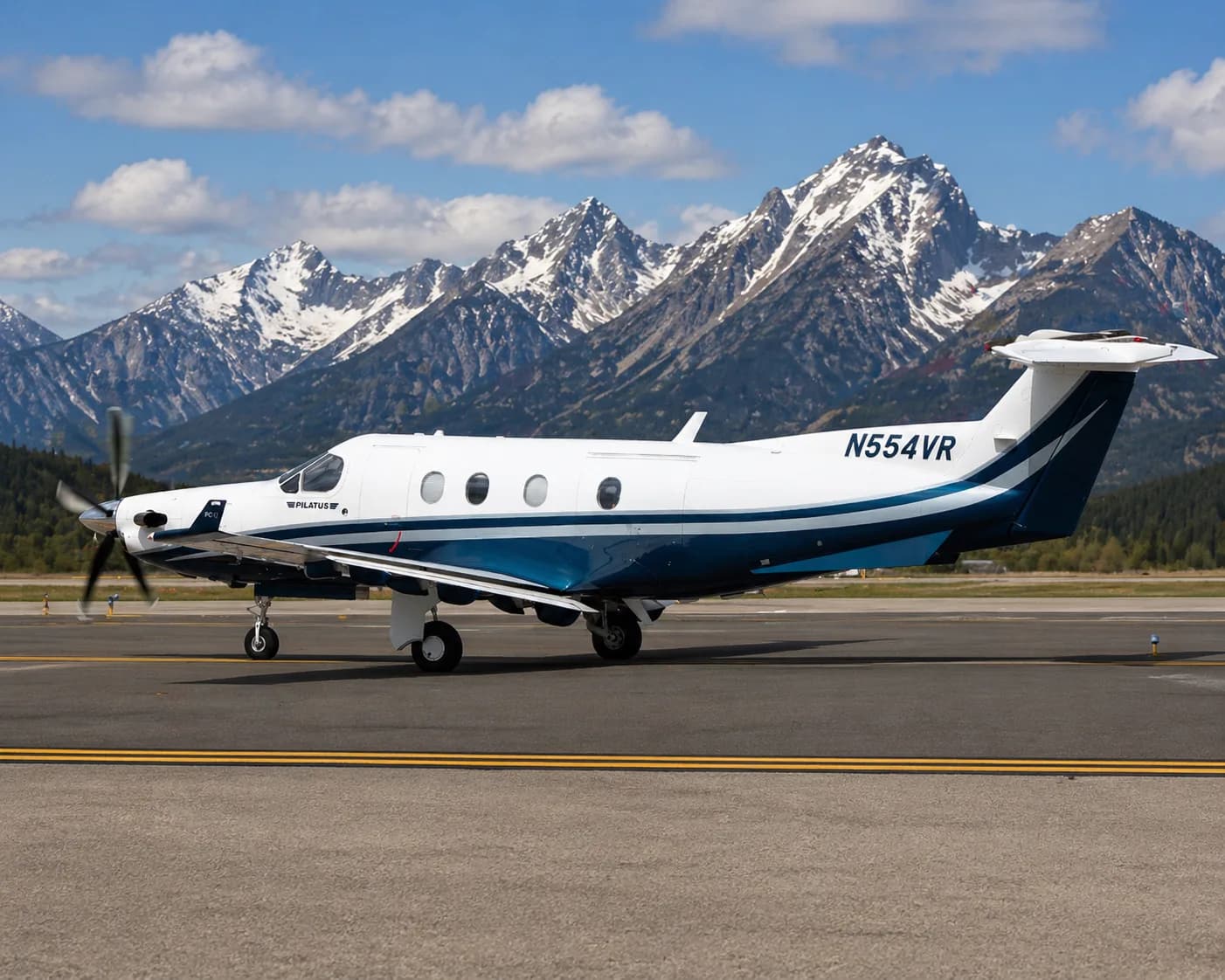 A Pilatus PC-12 aircraft with white and blue livery on a tarmac with snow-capped mountains in the background.