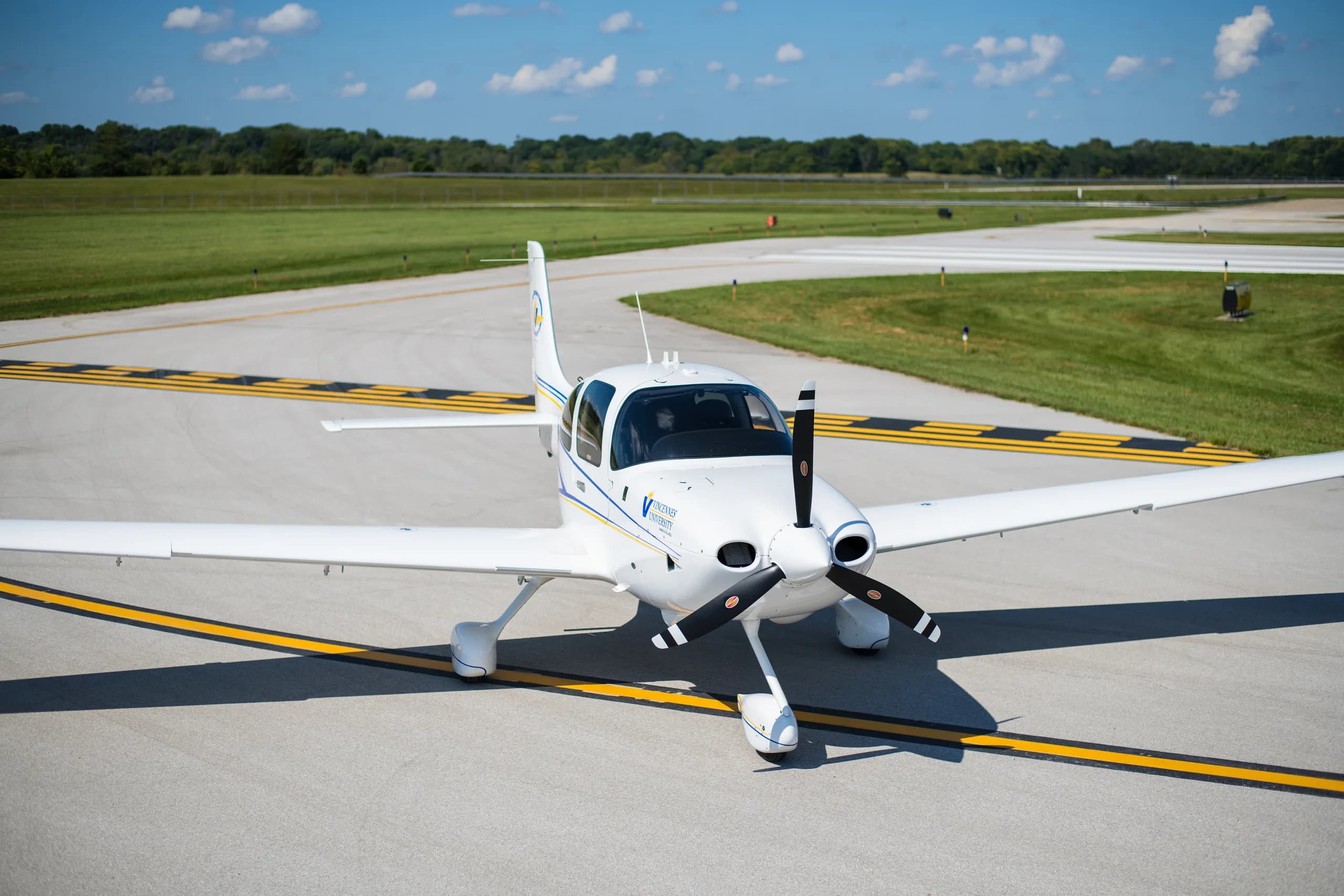 A white single-engine propeller aircraft with blue and yellow stripes and "Vincennes University" logo, parked on an asphalt runway with green fields and a clear blue sky in the background.