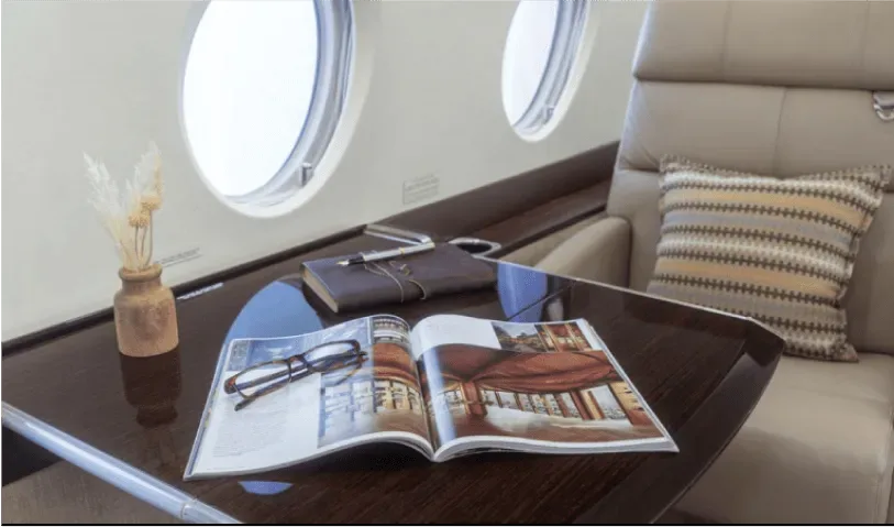 A close-up of a polished wooden table inside the aircraft cabin, displaying a magazine, reading glasses, and a notebook.