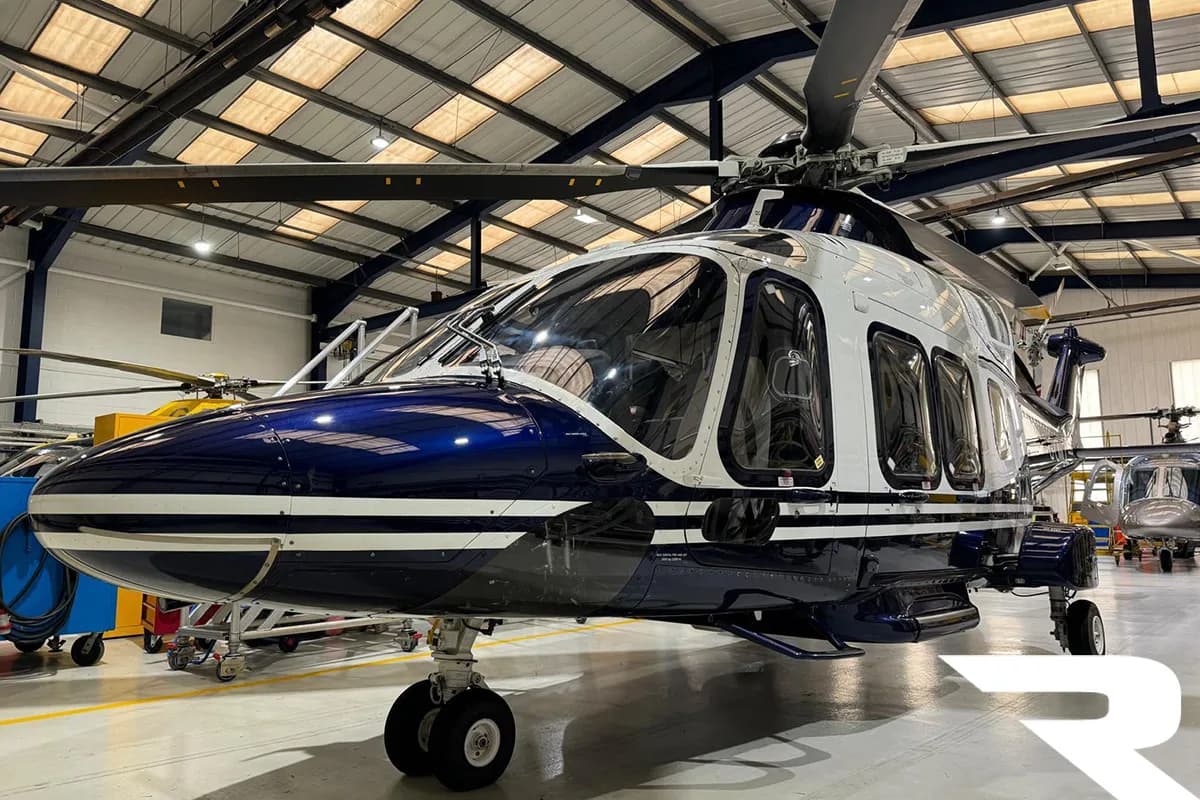 Front-left view of a blue and white helicopter with landing gear in a hangar, with other helicopters in the background.