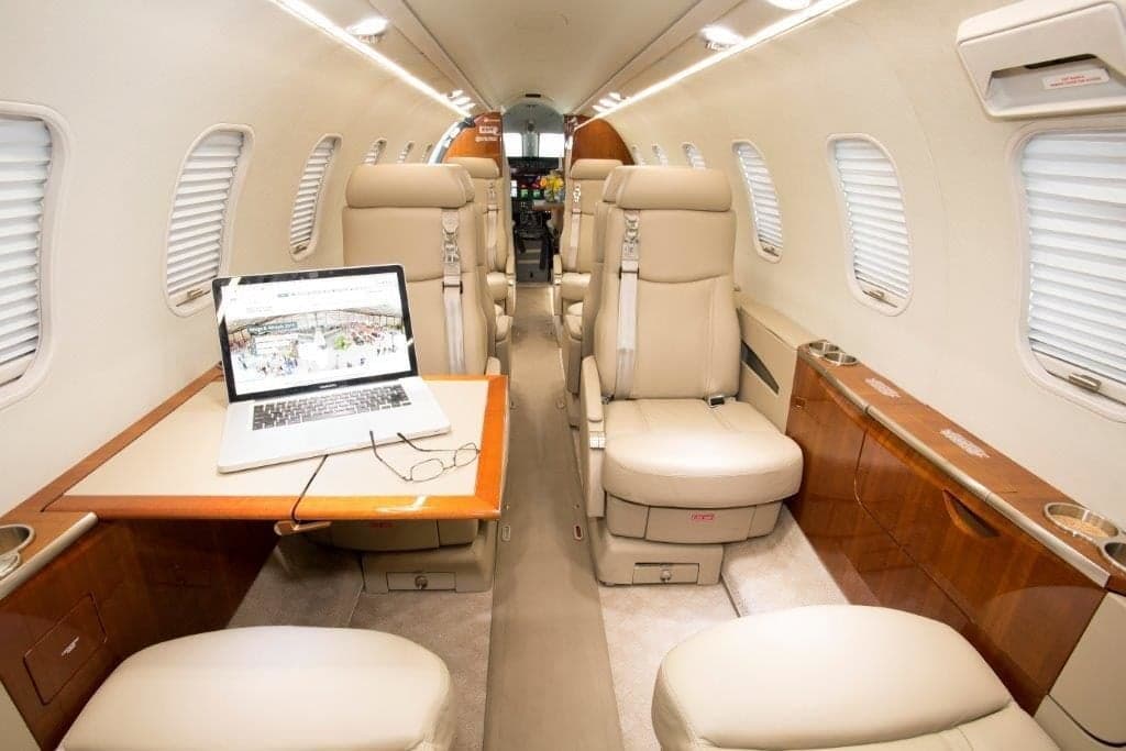 A view down the aisle of a private jet cabin with beige leather seats, wood trim, and a laptop on a table.