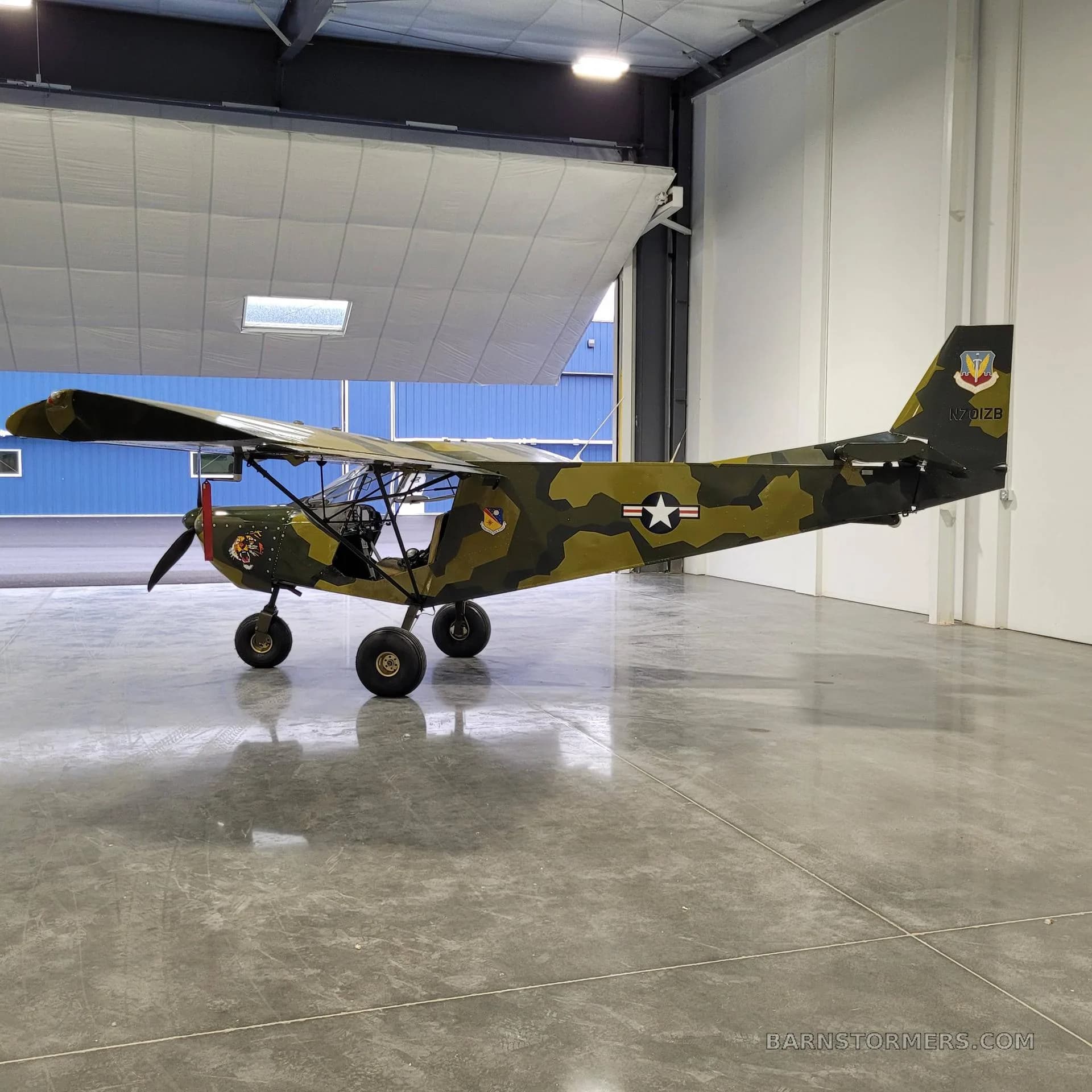 A full side view of a camouflage-painted, high-wing aircraft with a tiger decal and US Air Force markings, positioned in a hangar with an open door.