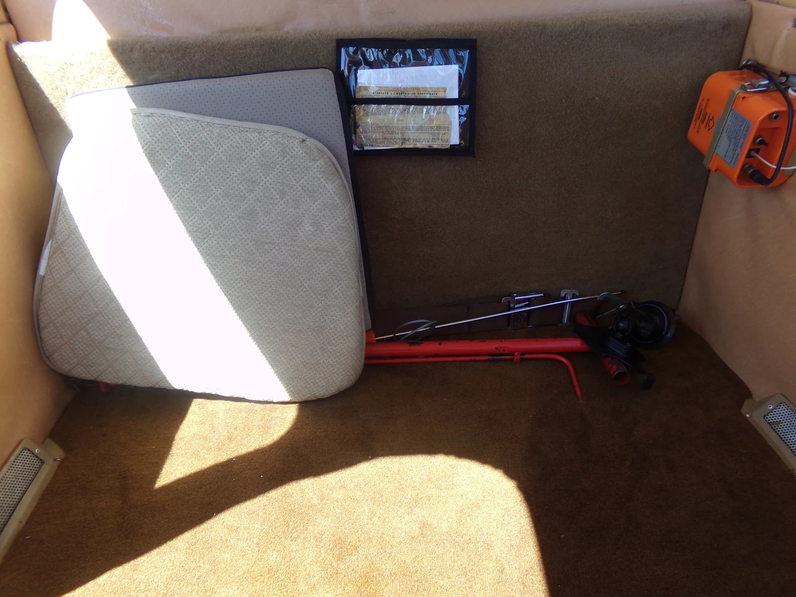 The rear baggage area of a small aircraft, showing a folded seat cushion, an emergency locator transmitter, and tools.