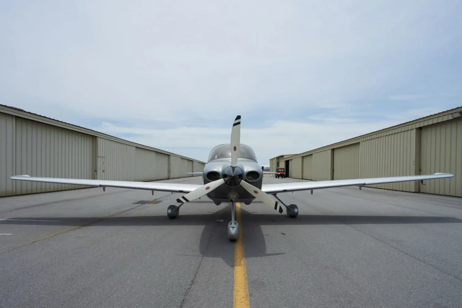 Front view of a silver Cirrus SR22 aircraft with a three-bladed propeller, parked between two hangars.