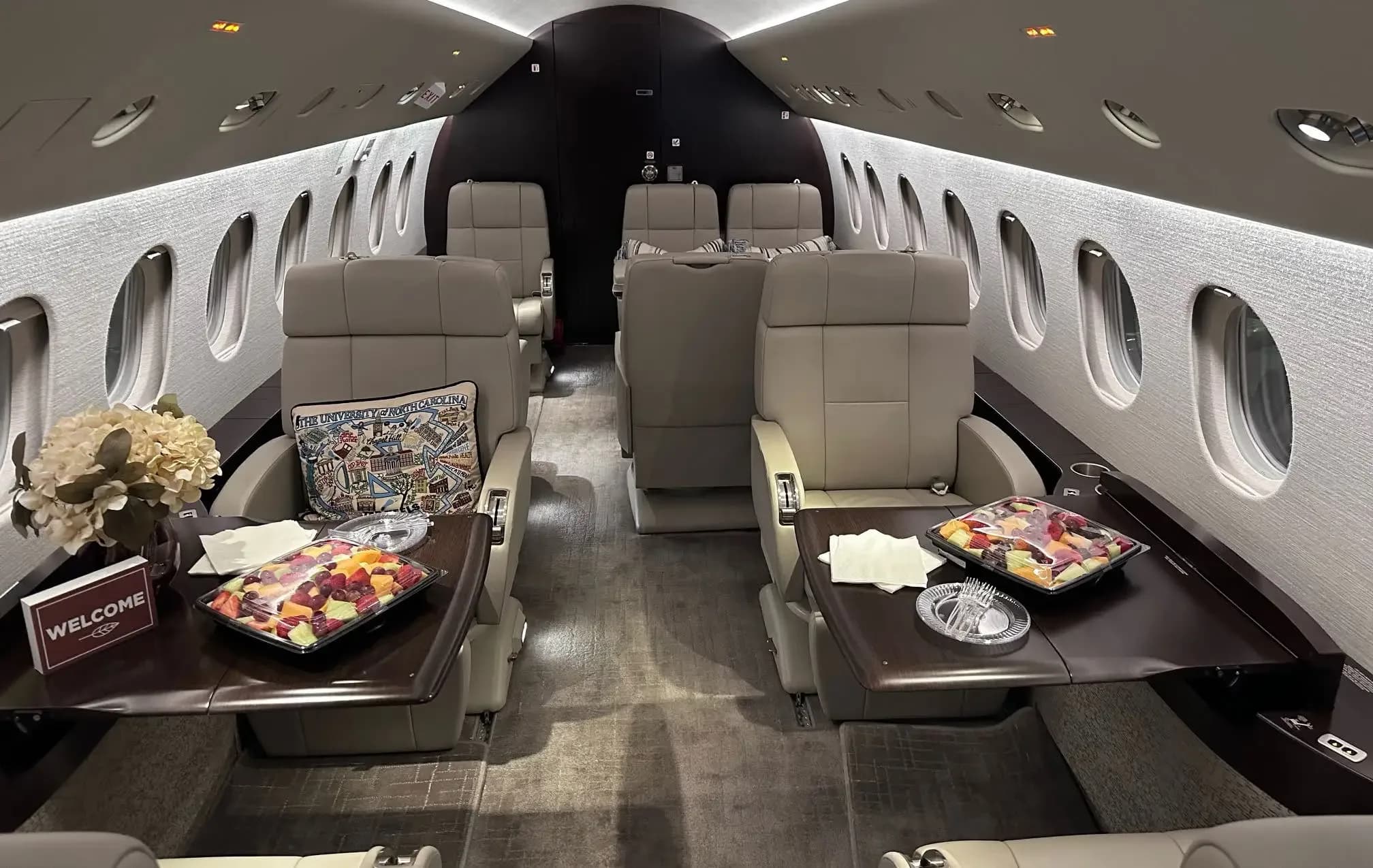The luxurious cabin interior of a business jet, featuring beige leather seats, dark wood tables with fruit platters, and a "WELCOME" sign.