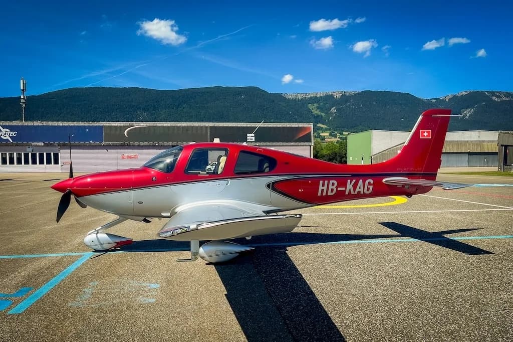 Red and silver Cirrus SR22 aircraft parked on an airfield, showing its side profile against a backdrop of mountains and buildings.