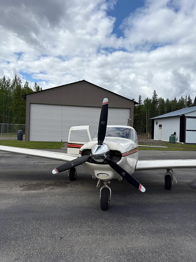 Front view of a white and brown aircraft with its door open, parked in front of a hangar.