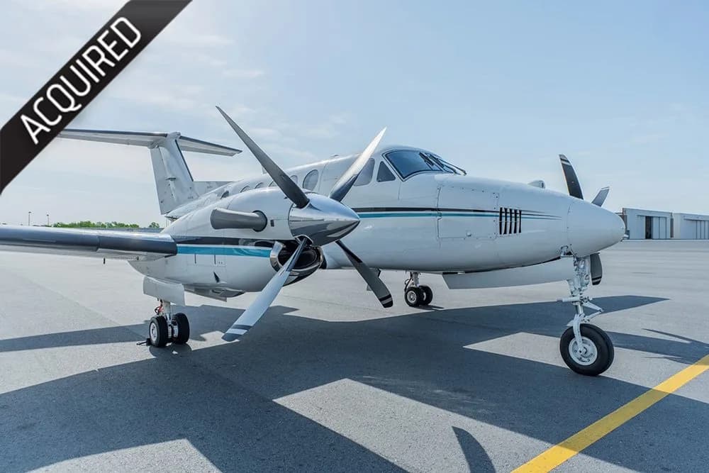 A white and blue twin-turboprop aircraft, likely a King Air, parked on an airport tarmac with an 'ACQUIRED' banner.