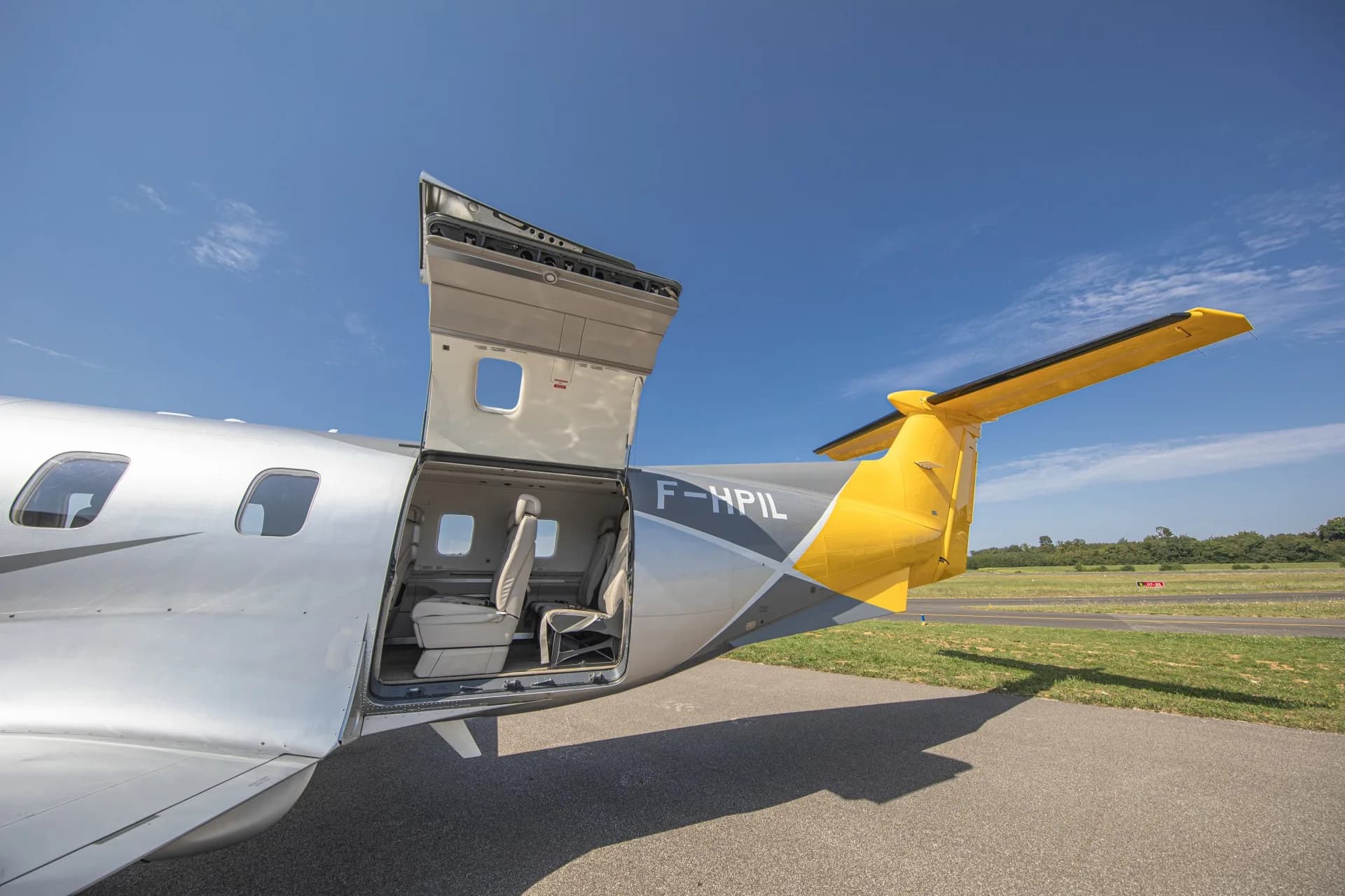 The rear cargo door and passenger door of the silver and yellow Pilatus PC-12 aircraft (F-HPIL) are open on the tarmac.