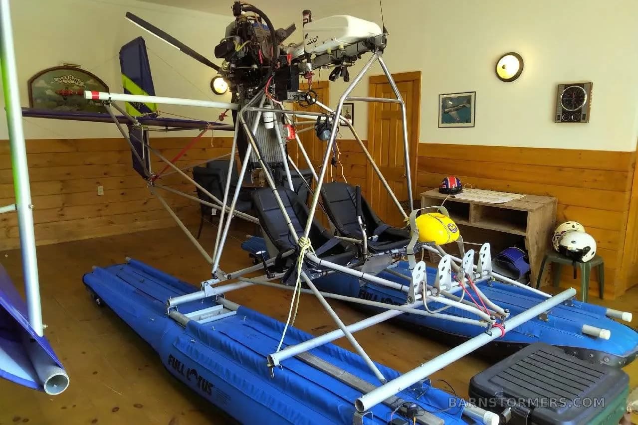 A blue ultralight seaplane with exposed frame and engine, stored indoors in a room with wood paneling.