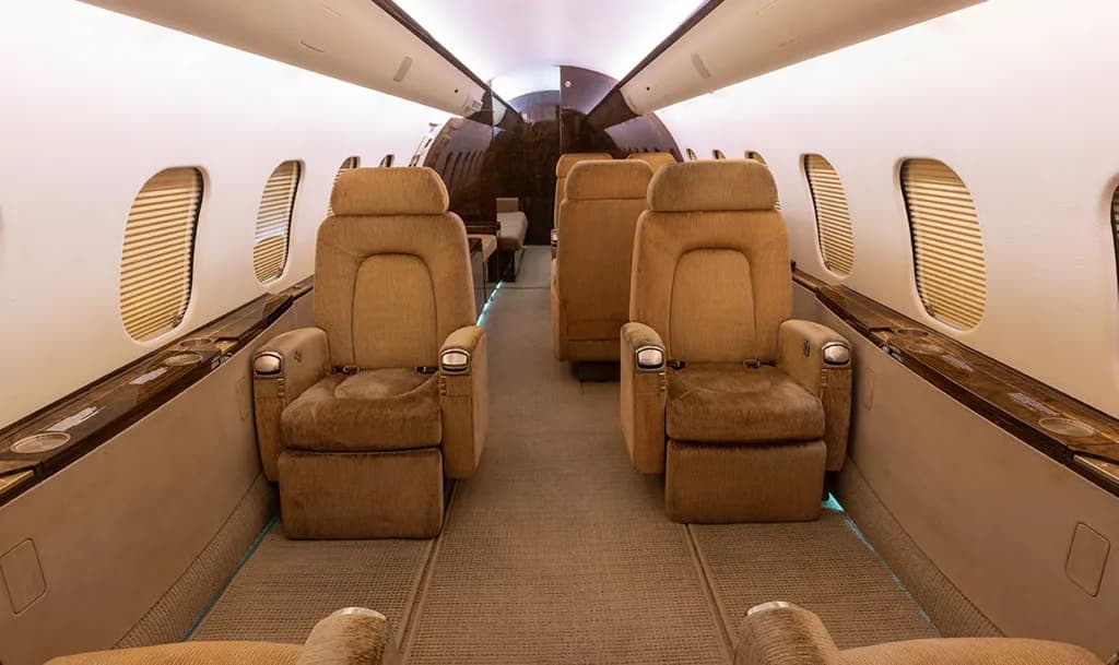 The luxurious interior cabin of a private jet, featuring two rows of plush tan individual seats and dark wood accents.