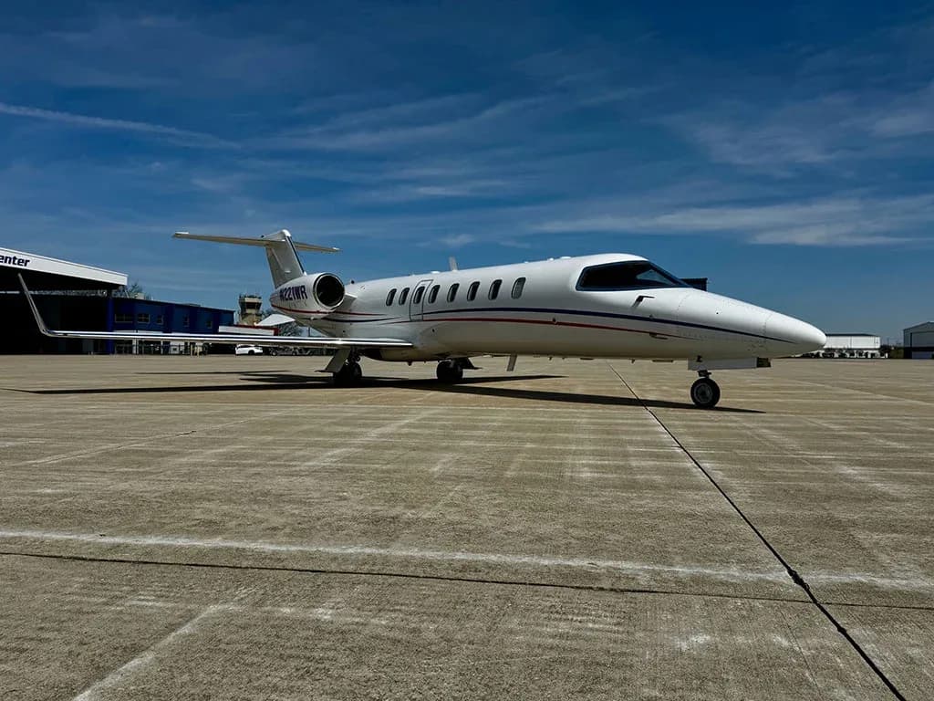 A white Learjet 60, registration N221WP, with red and blue stripes, is parked on an airport tarmac under a blue sky.