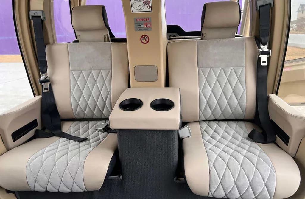 The rear cabin interior of a helicopter, showing two beige and gray quilted seats with a central armrest and cup holders.