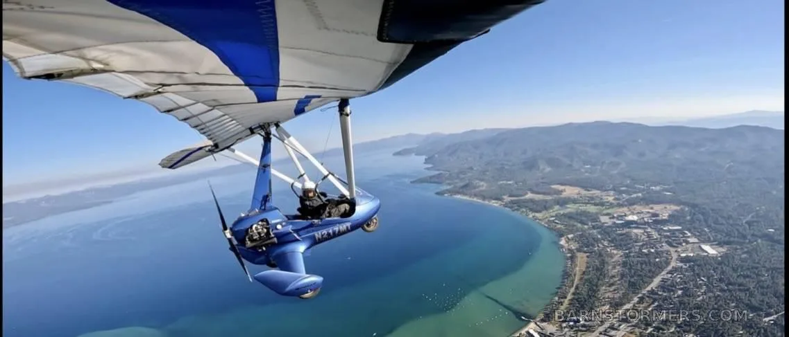 A blue ultralight aircraft with registration N217MT flies over a large blue lake and a forested coastline.