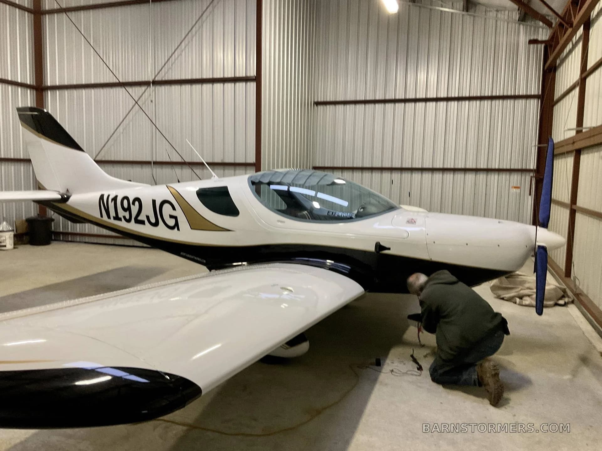 A white and black aircraft with registration N192JG parked in a hangar, with a person working near the propeller.
