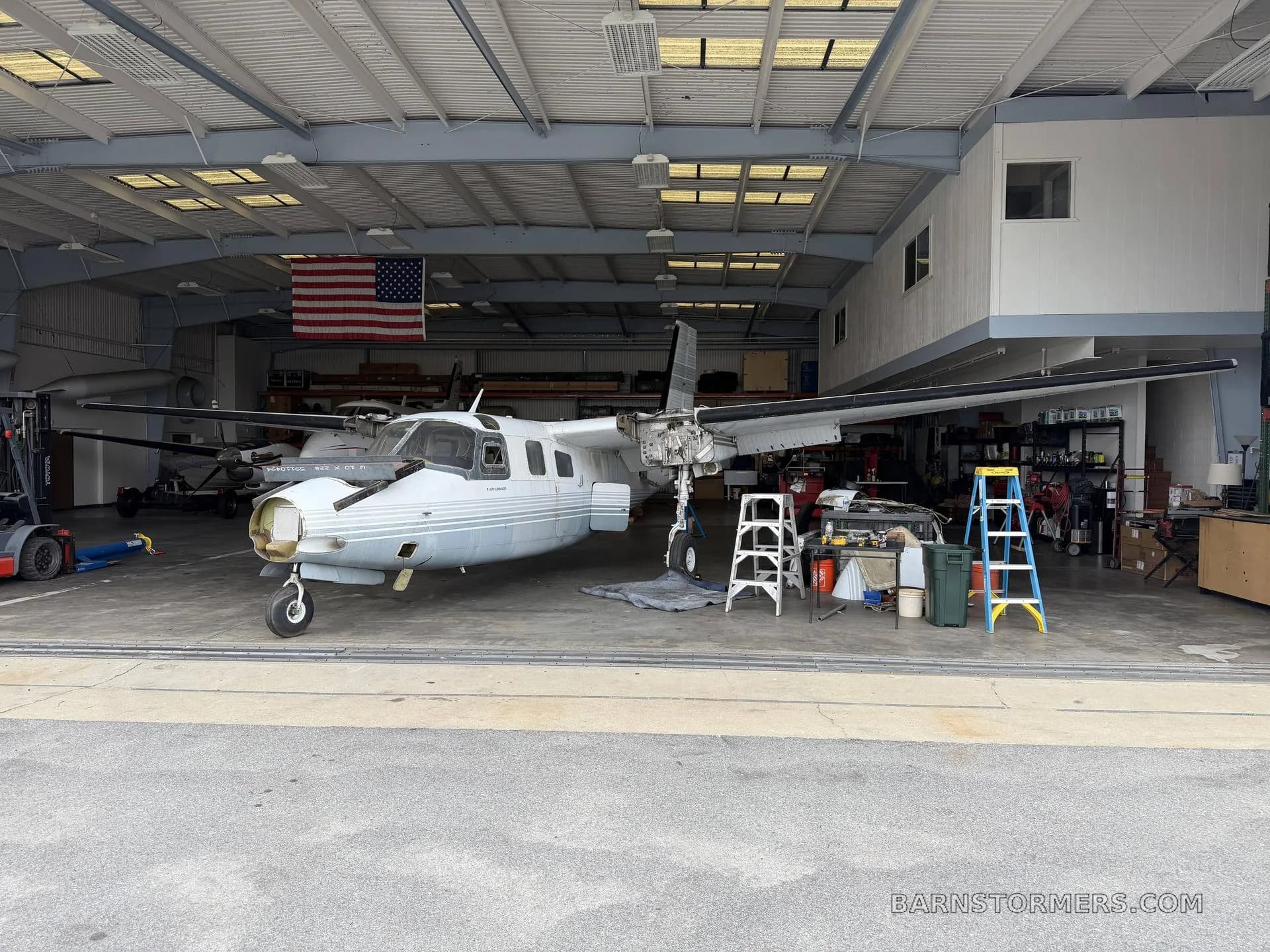 A white twin-engine aircraft with its nose cone and engine cowlings removed, parked in a hangar with tools and an American flag.