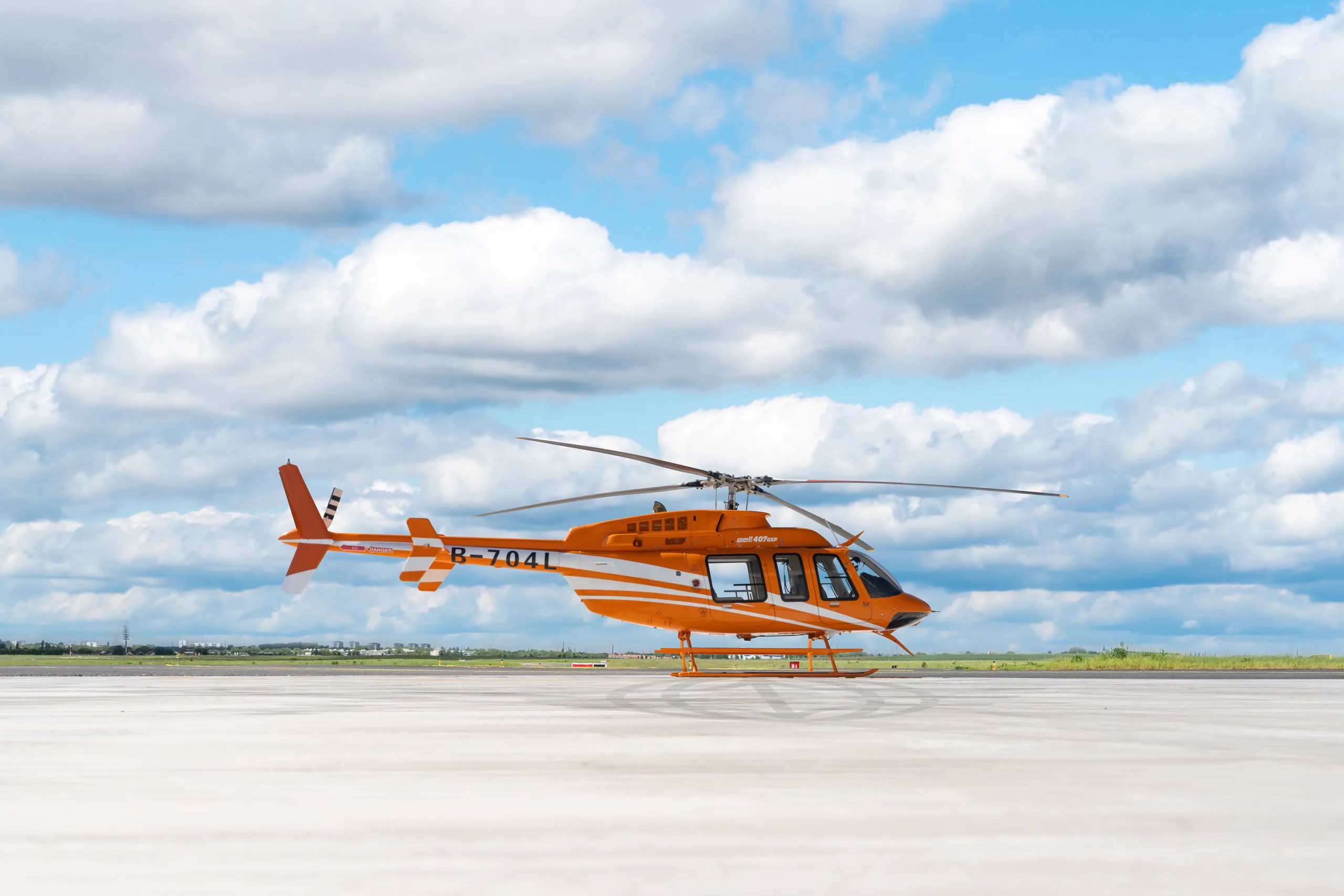 An orange and white Bell 407GXi helicopter is parked on the tarmac under a cloudy sky.