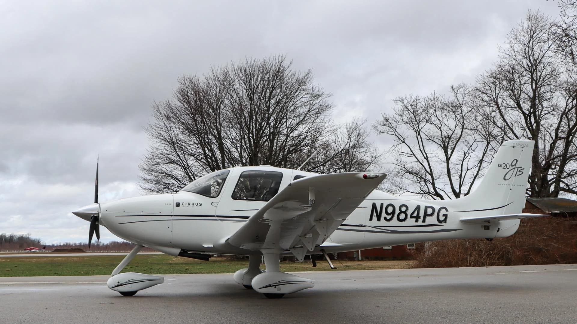 A white Cirrus SR20 GTS aircraft, N984PG, is parked with its right side visible against a backdrop of bare trees.