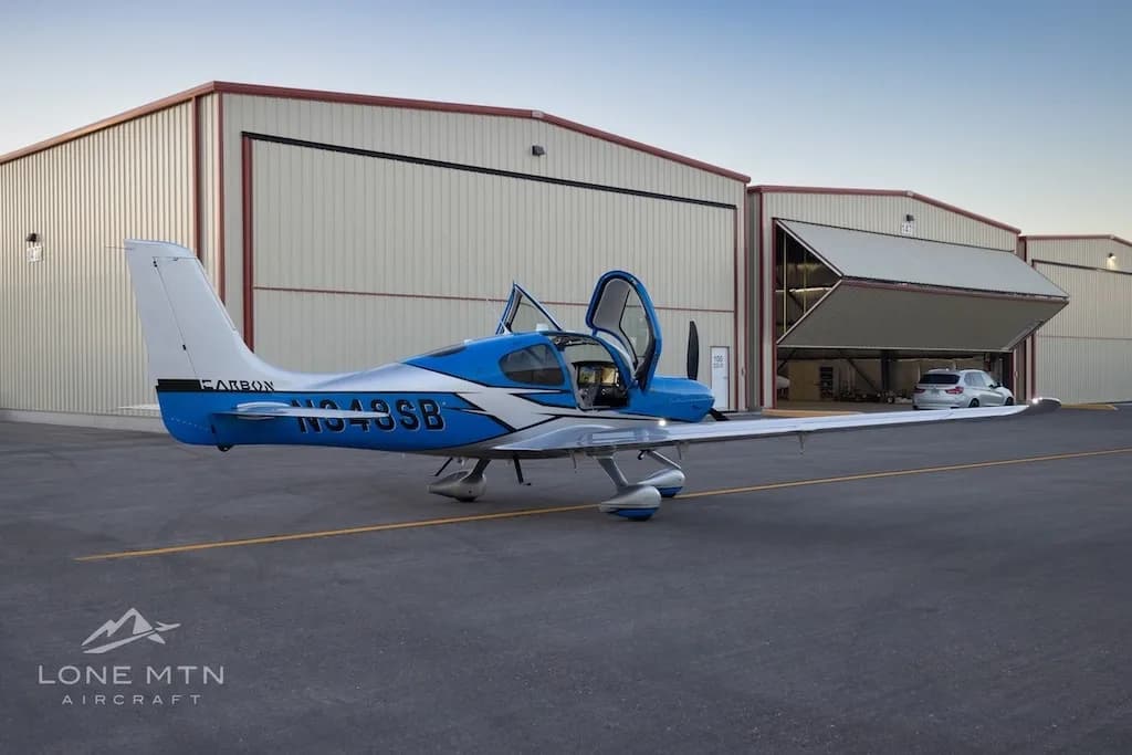 A blue and silver Cirrus SR22 aircraft with open doors parked on an airfield, with a hangar door opening in the background.