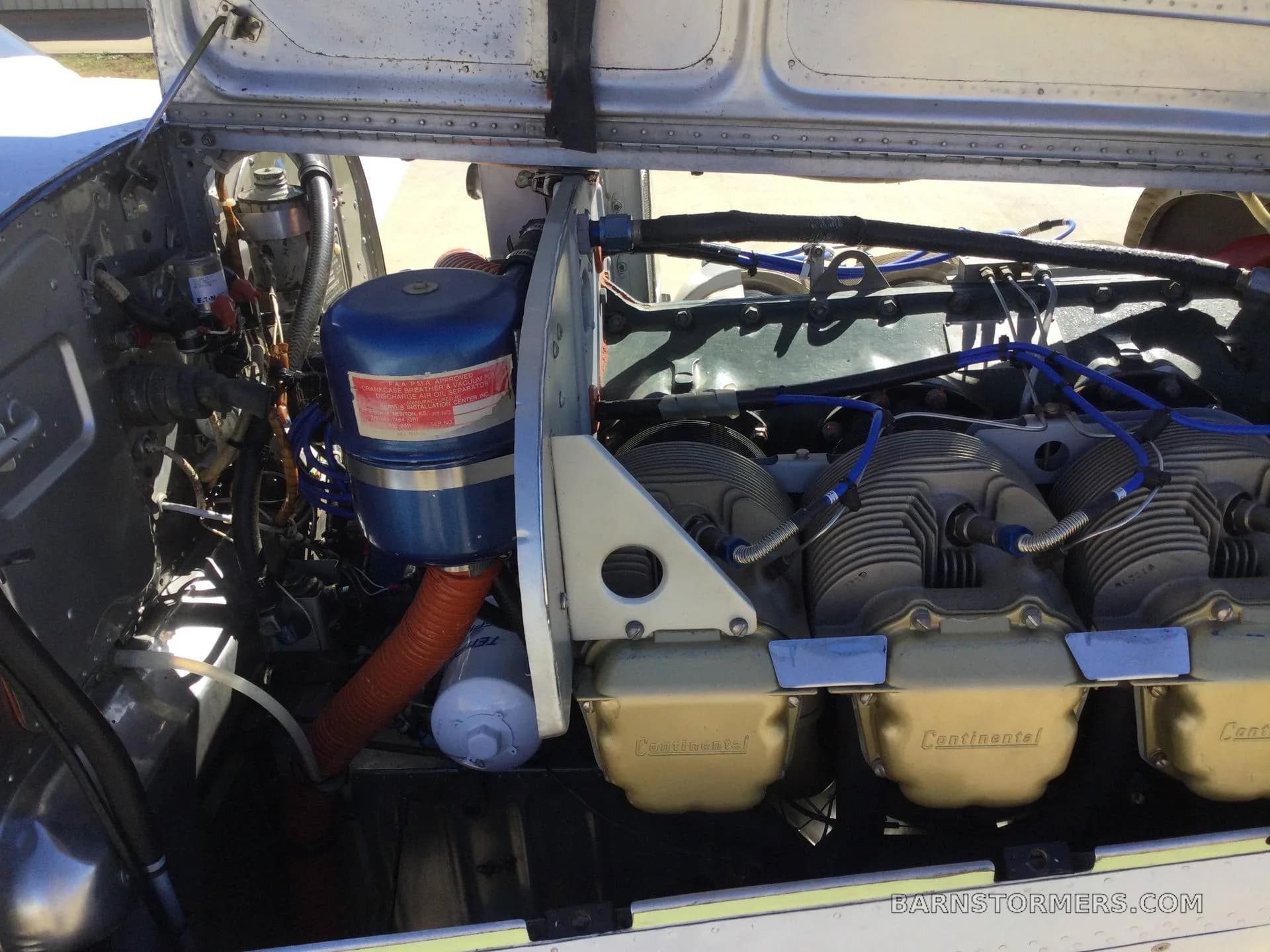A close-up view of an aircraft engine compartment showing a blue air filter, gold Continental valve covers, and various hoses and wires.