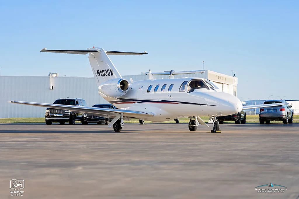 A front-side view of a white Cessna Citation CJ3 (N903GW) with blue and red stripes parked on an airfield.