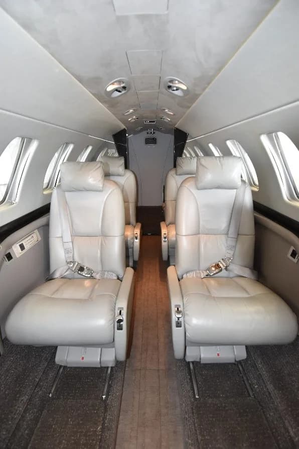 The interior cabin of a private jet, showing multiple rows of light gray leather seats and a central aisle.