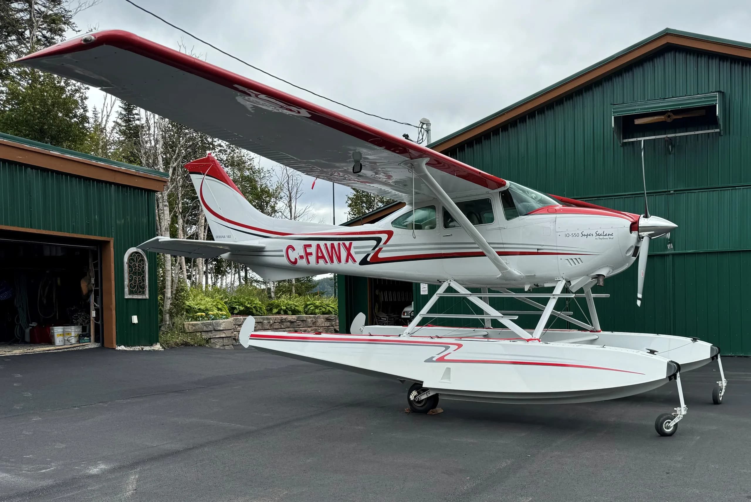 A white and red Cessna 182 seaplane with registration C-FAWX is parked on asphalt next to a green building.