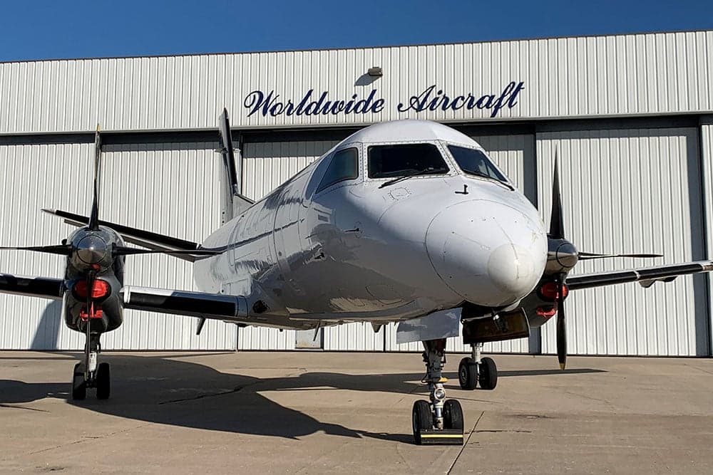 Front view of a white Beechcraft 1900C aircraft parked in front of a hangar.