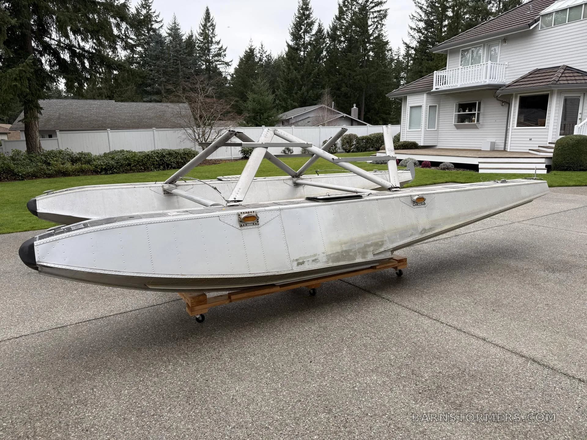 A pair of aluminum aircraft floats with mounting struts, viewed from the rear-side, on a concrete driveway with trees in the background.
