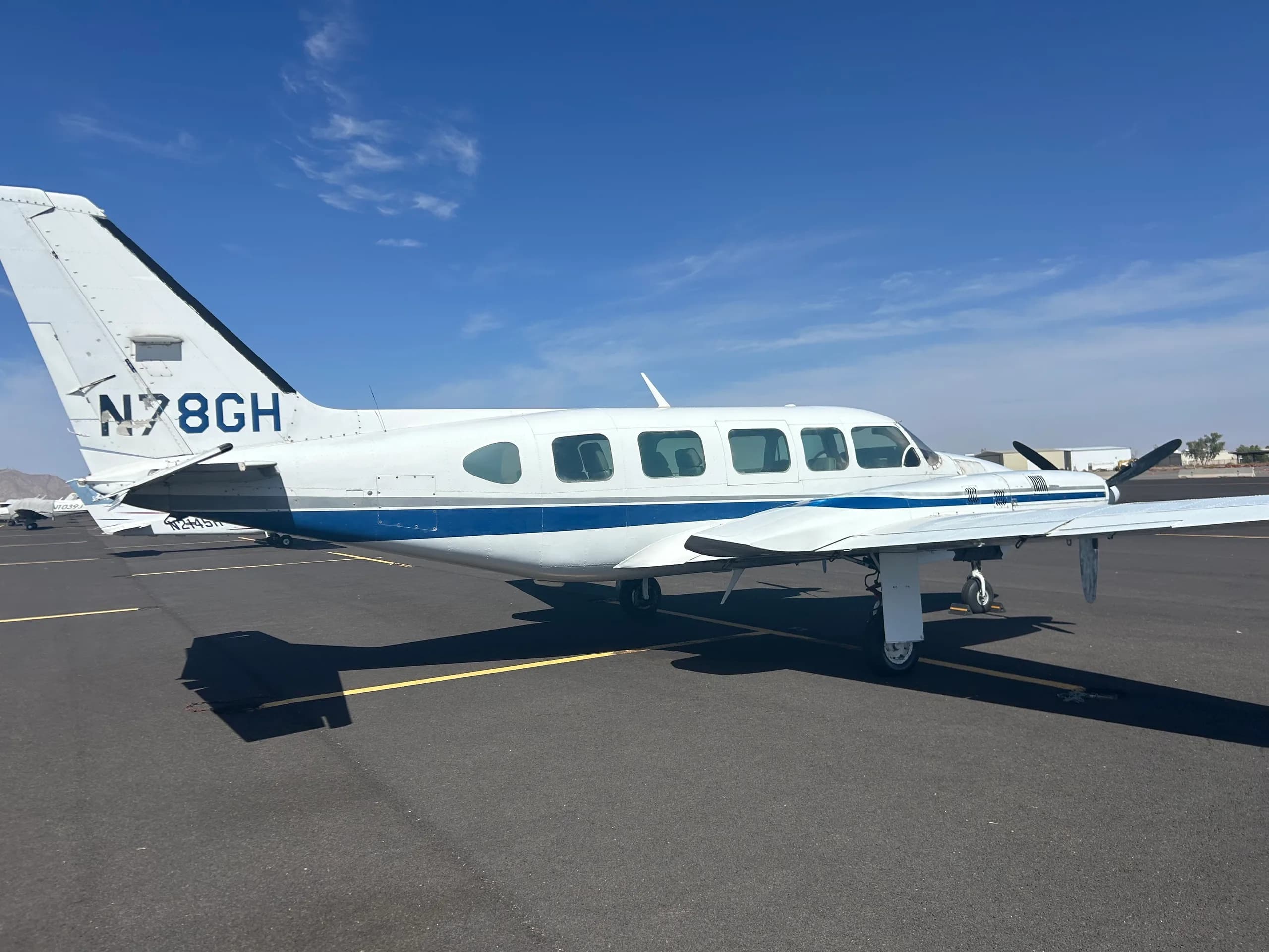 A white twin-engine aircraft with blue and grey stripes, registration N78GH, parked on an asphalt tarmac under a clear blue sky.