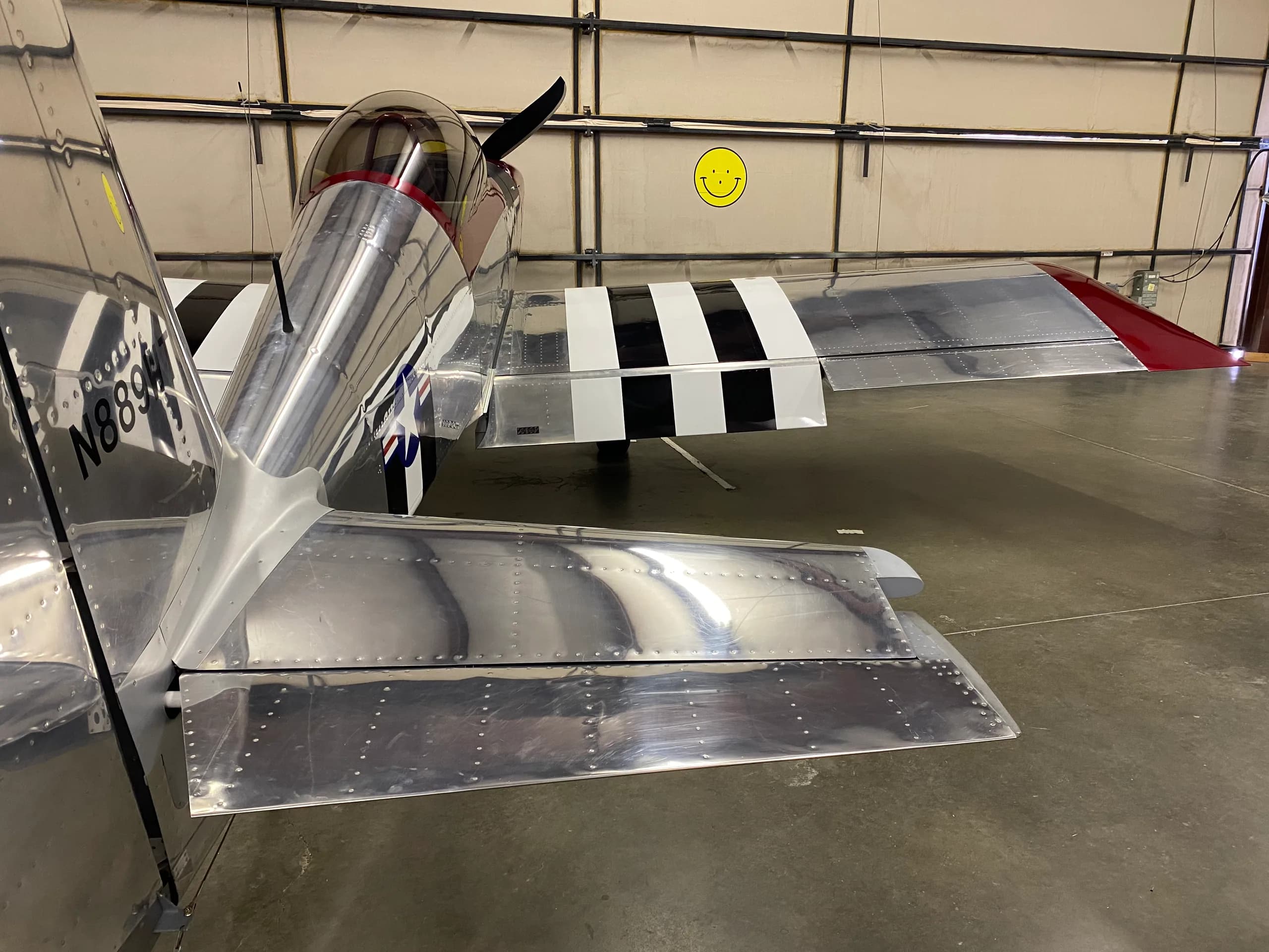Rear view of a silver and red Vans RV-8 aircraft with black and white invasion stripes on the wing, parked inside a hangar.