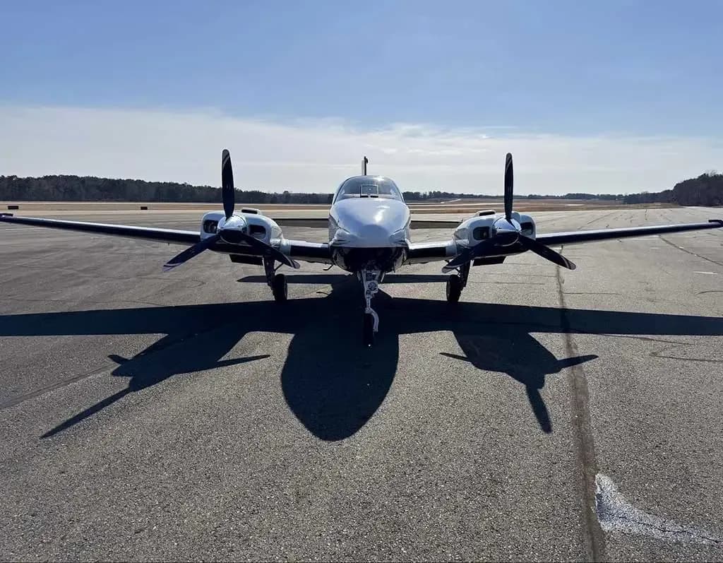 A white twin-engine aircraft, likely a Beechcraft Baron, parked on an airfield, viewed from the front.