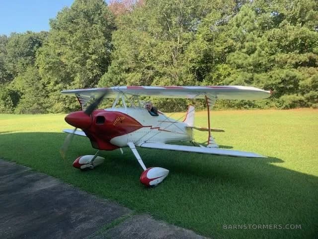 A red and white 'Little Toot' biplane with a pilot in the cockpit, parked on a grassy field next to a paved path.