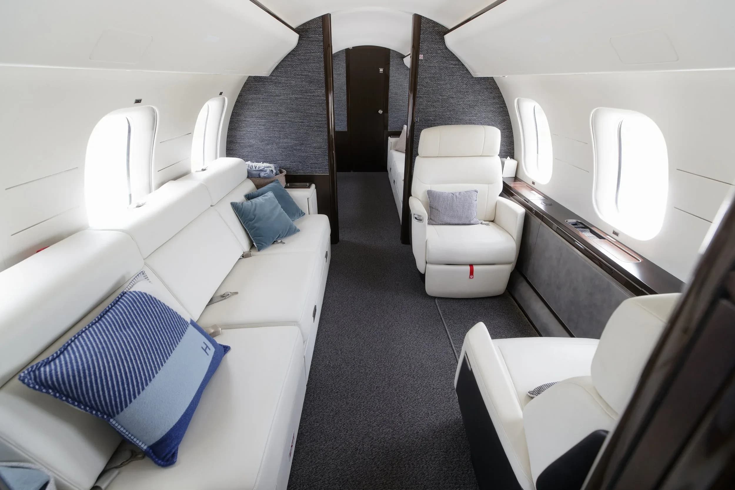 The aft cabin of a private jet, featuring a long white sofa with blue pillows and an individual white leather seat.