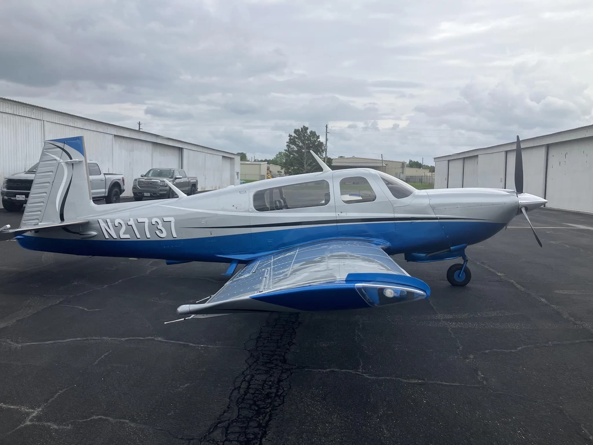 A silver and blue single-engine propeller aircraft, registration N21737, parked on an asphalt apron with hangars in the background under a cloudy sky.