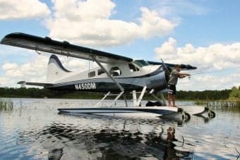 A white and silver float plane takes off from a lake with mountains in the background.
