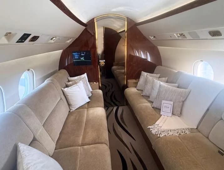 A private jet cabin interior featuring two long, comfortable beige couches with white pillows, facing each other.