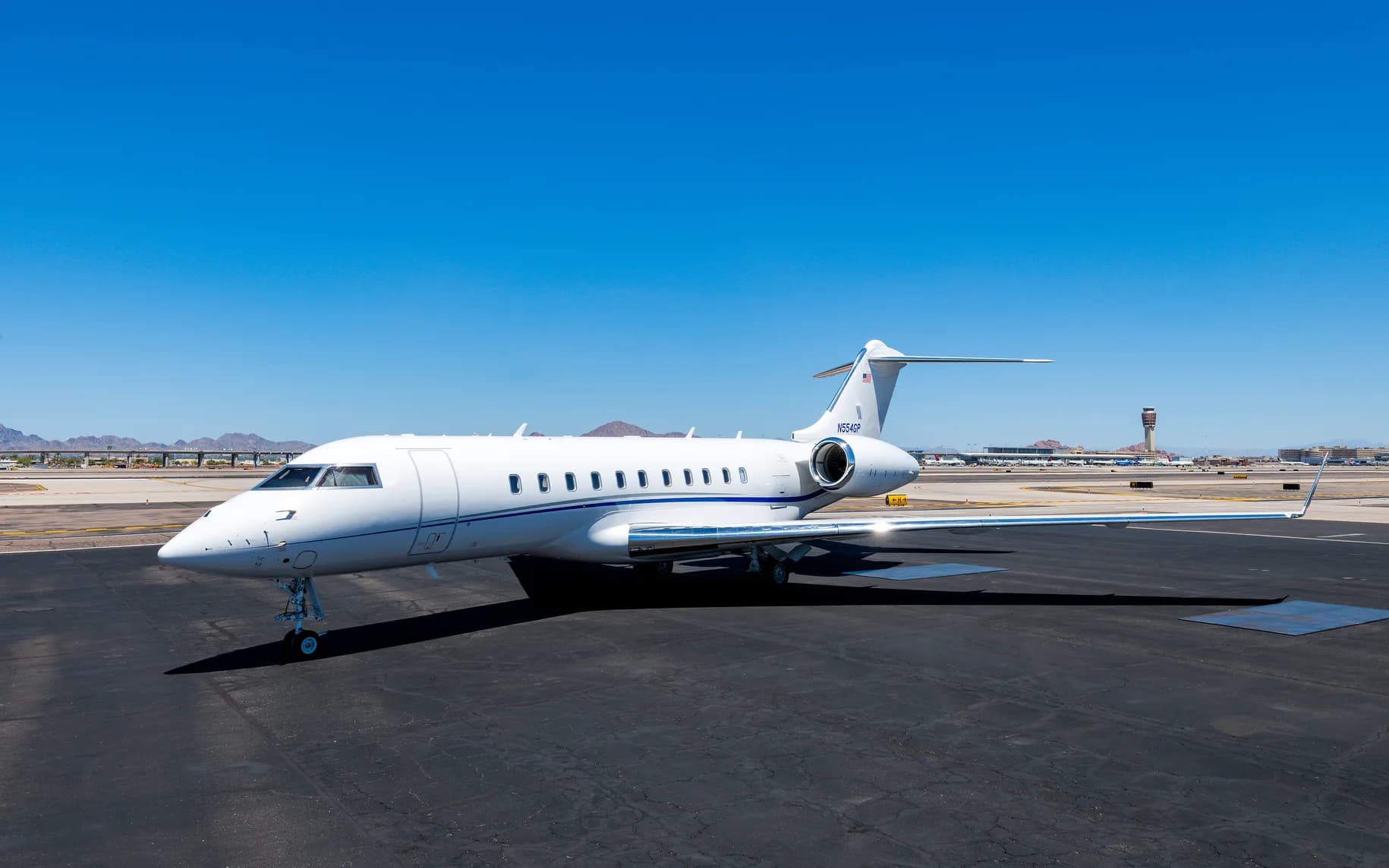A white private jet with a blue stripe is parked on an airport tarmac on a sunny day, with mountains and a control tower in the background.