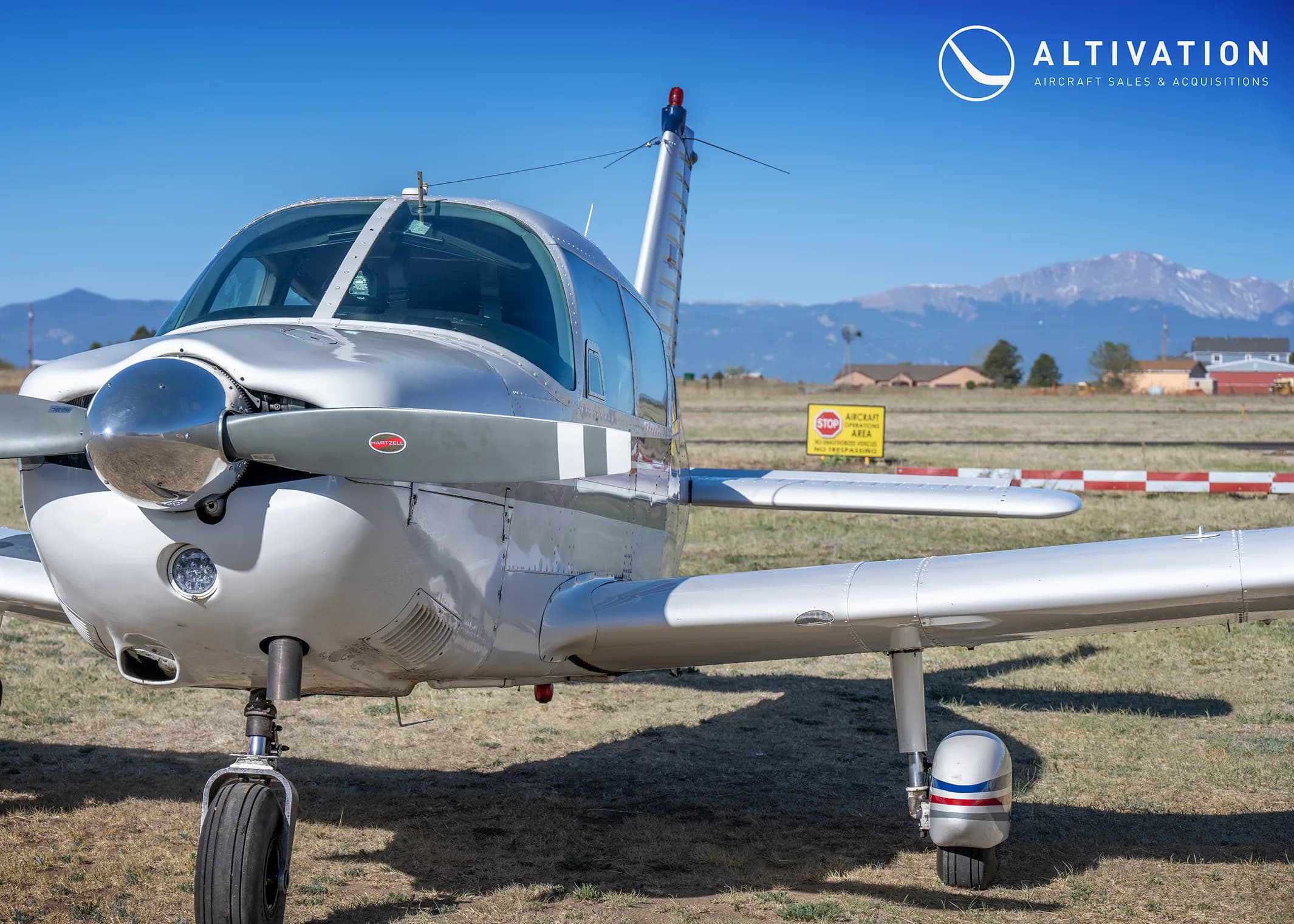 A close-up front view of a white Piper Cherokee aircraft with mountains in the background.